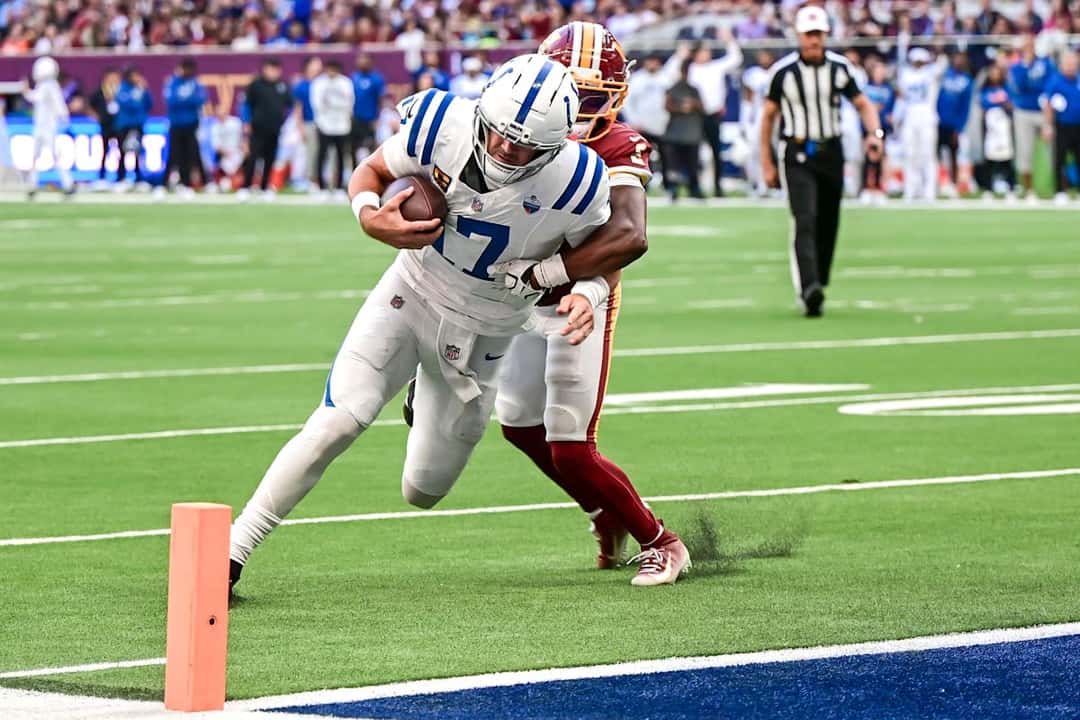 Daniel Jones, quarterback de los Colts, se lanza hacia la zona de anotación marcado por un defensor de los Commanders