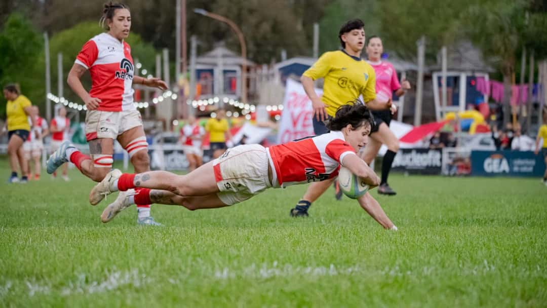 Una jugadora de camiseta roja y blanca se zambulle para apoyar un try en el Nacional de Clubes Femenino, con la árbitra cerca