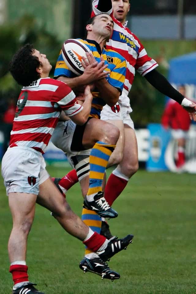 Foto de archivo: un jugador de Hindú, con camiseta a rayas azules y amarillas, salta a buscar la pelota en el aire entre dos rivales de Alumni con camisetas a rayas rojas y blancas