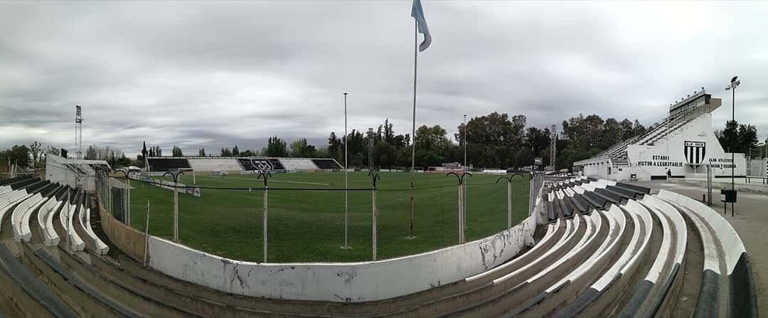 Vista panorámica lateral del estadio Víctor Antonio Legrotaglie, de Gimnasia y Esgrima de Mendoza, con las gradas blancas y negras y el escudo del club sobre la tribuna (foto de archivo de 2017: Iván Aguilar, Wikimedia Commons, CC BY-SA 4.0)