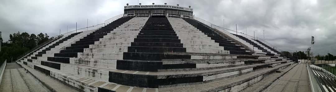 La platea oeste del estadio Víctor Antonio Legrotaglie, de Gimnasia y Esgrima de Mendoza, con sus gradas pintadas de blanco y negro y el nombre del club en lo alto; foto de archivo de 2017 (foto: Iván Aguilar / Wikimedia Commons, CC BY-SA 4.0)