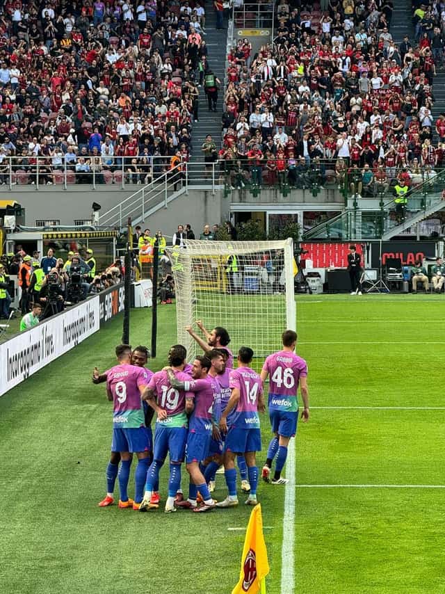 Jugadores del Milan, con una camiseta alternativa rosa y azul, se abrazan para festejar un gol junto al córner, con la tribuna de San Siro llena detrás; foto de archivo de abril de 2024 (foto: Alexdevil, Wikimedia Commons, dominio público, CC0)