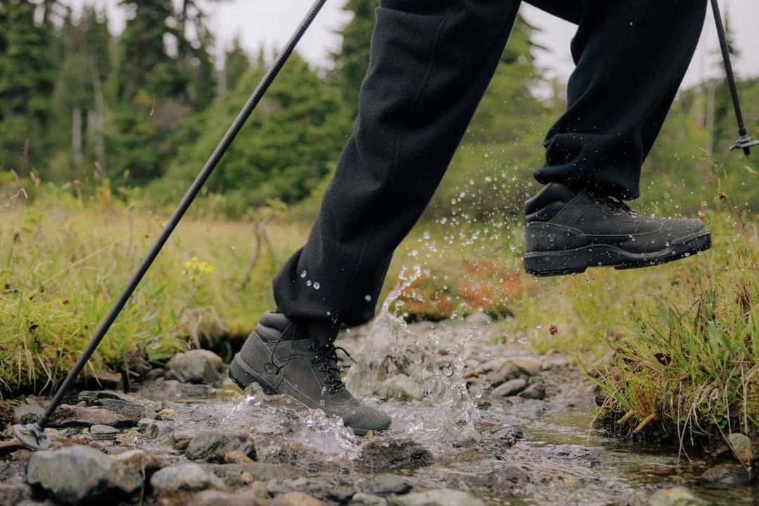 Persona cruzando un arroyo de montaña con la HAVEN x Timberland Field Boot puesta y bastones de trekking, en la campaña de la colaboración