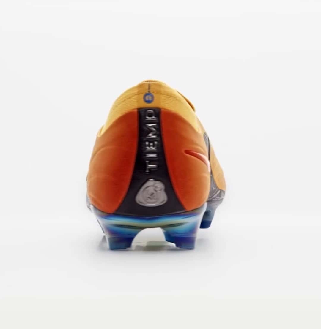 Talón del botín Kids of Immigrants x Nike Tiempo Maestro Elite Sundial sobre fondo blanco, en naranja y rojo, con una franja negra vertical que dice Tiempo y la base de tapones en tonos azules