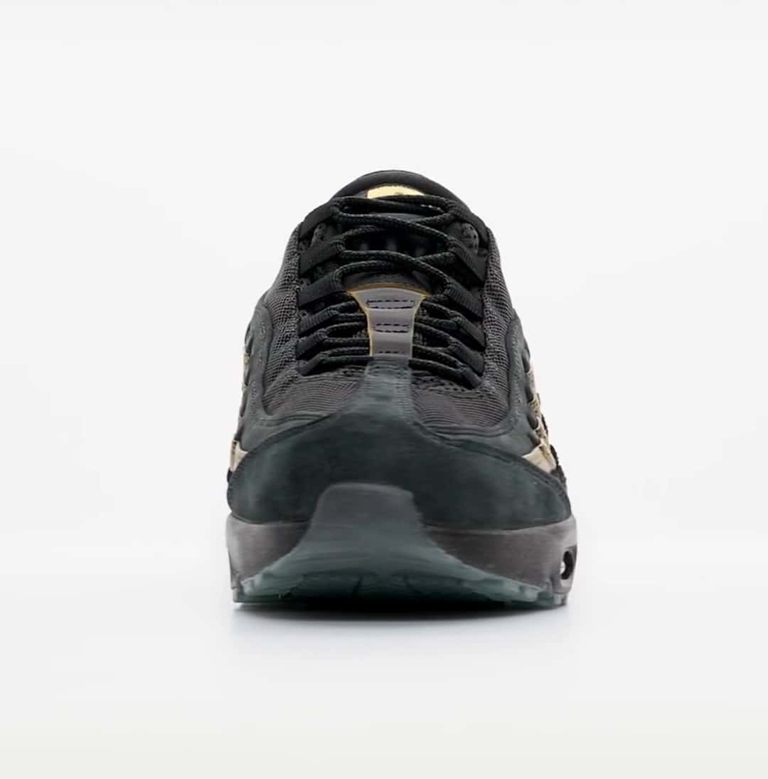 Kyle Wilson x Nike SB Air Max 95 visto de frente sobre fondo gris claro, con la puntera de gamuza negra, los cordones negros y los paneles con brillo dorado