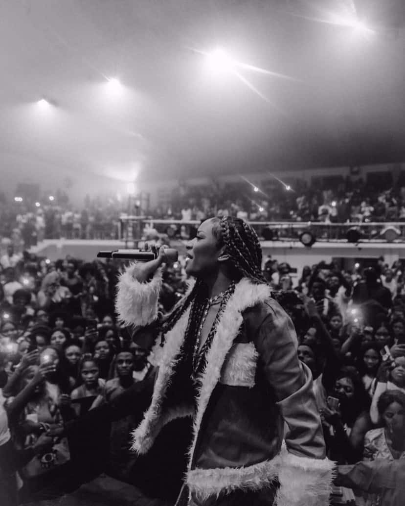 Ayra Starr canta con el micrófono en la mano, con trenzas largas y un abrigo con piel blanca, frente a un público con los celulares en alto, en un show en la Universidad de Benín, Nigeria, en 2021, en blanco y negro (foto de archivo). Foto: Tomisin Afolabi, CC BY 2.0, vía Wikimedia Commons