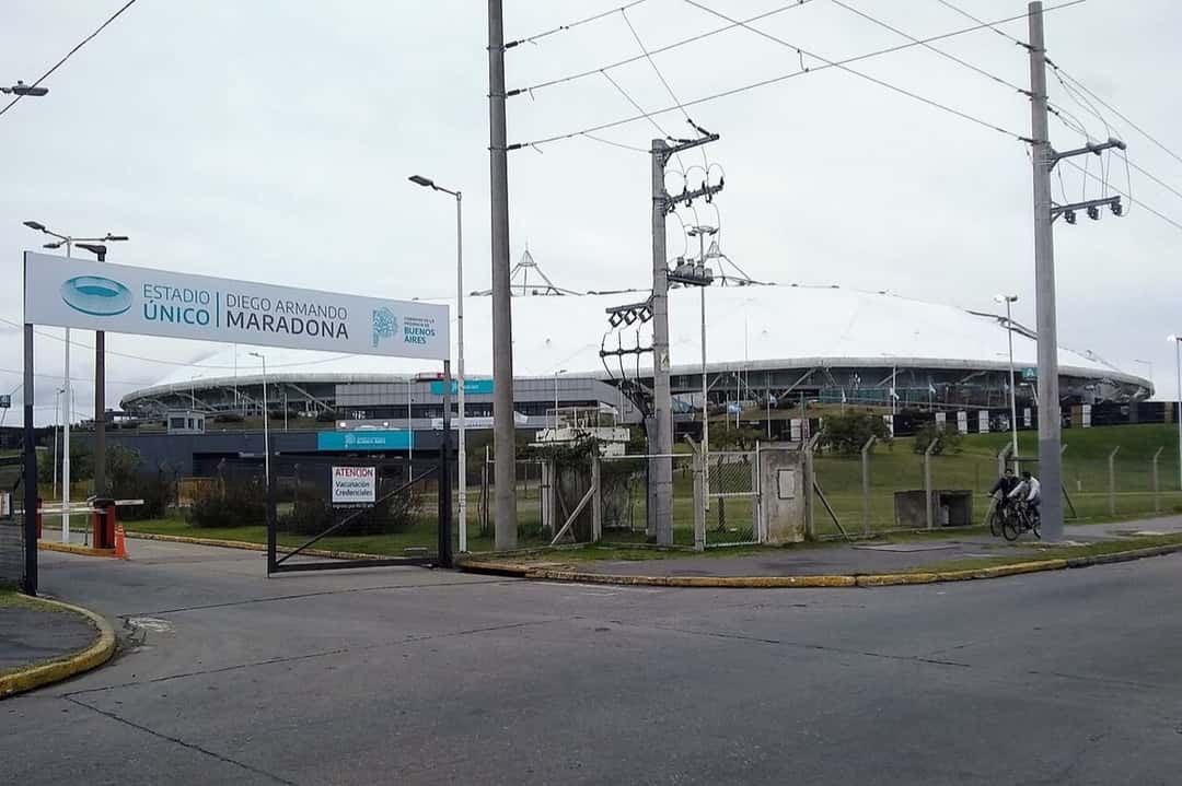 Acceso al Estadio Único Diego Armando Maradona de La Plata, con el cartel del estadio y el techo blanco detrás de la reja. Foto: Cazadoroculto, CC BY-SA 4.0, vía Wikimedia Commons