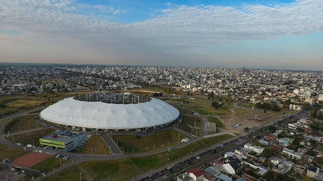 Vista aérea del Estadio Único de La Plata, con su techo blanco, el predio verde alrededor y la ciudad de fondo. Foto: Iván Furlan, CC BY-SA 4.0, vía Wikimedia Commons