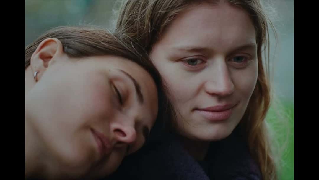 Fotograma del video oficial de «blue»: dos jóvenes abrazadas, una con los ojos cerrados apoyada en el hombro de la otra, sobre un fondo verde
