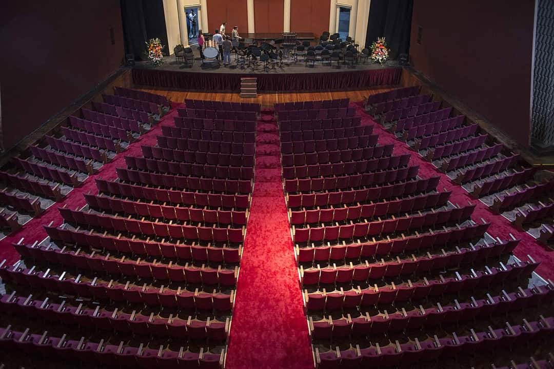 Sala del Teatro Coliseo de Buenos Aires vista desde arriba: filas de butacas bordó a ambos lados de un pasillo central alfombrado en rojo, y al fondo el escenario con atriles, sillas y una batería. Foto: Enrico Fantoni, Wikimedia Commons, CC BY-SA 4.0