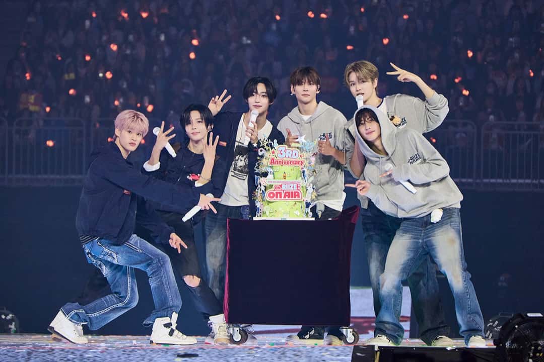 Foto oficial de archivo de RIIZE en el cierre de su fan meeting de tercer aniversario: los seis integrantes, con buzos grises, camperas y jeans, posan con micrófonos y haciendo la V con los dedos junto a una torta verde con el cartel 