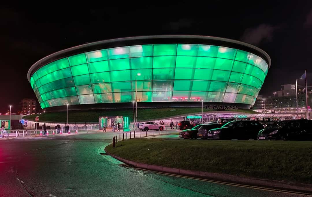 El OVO Hydro de Glasgow de noche, con su fachada redonda de vidrio iluminada de verde y autos estacionados enfrente (imagen del lugar donde arrancó la gira). Foto: Thomas Nugent, CC BY-SA 2.0, vía Wikimedia Commons