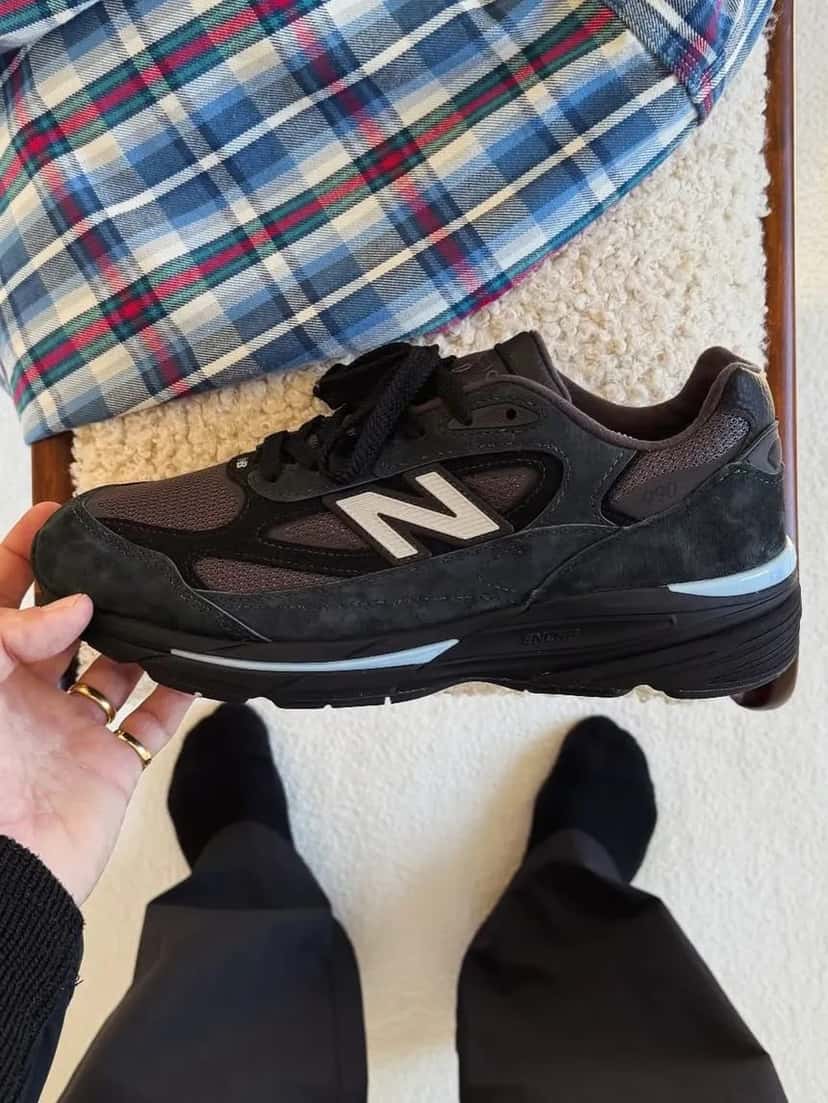 New Balance 990v7 Black sostenido en la mano, visto de costado y desde arriba, con los cordones negros, la N clara y los detalles celestes en la entresuela