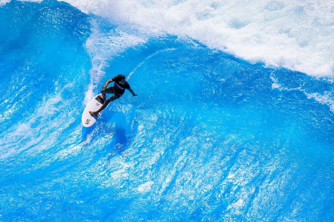 Un surfista con traje negro baja por la pared de la ola artificial de Wai Kai, con la espuma blanca rompiendo detrás
