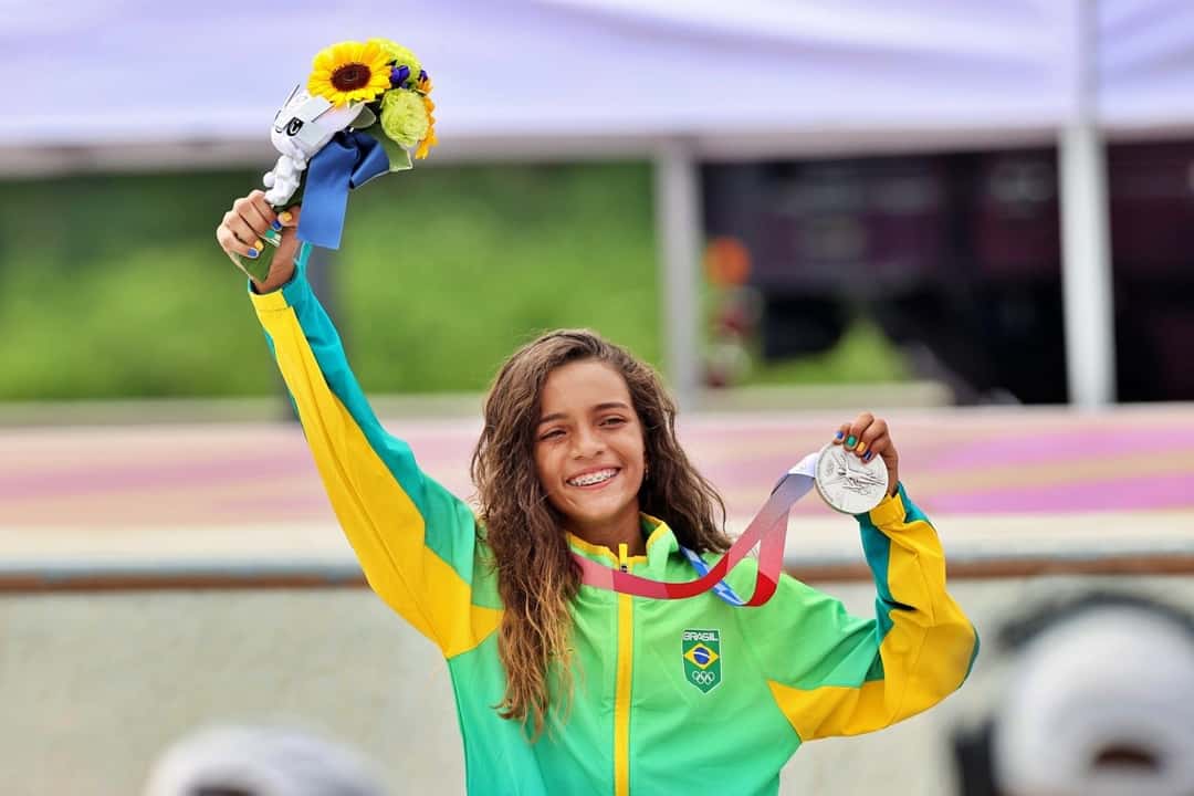 Rayssa Leal sonríe con el brazo en alto, un ramo de flores en una mano y la medalla de plata en la otra, con la campera verde y amarilla de Brasil