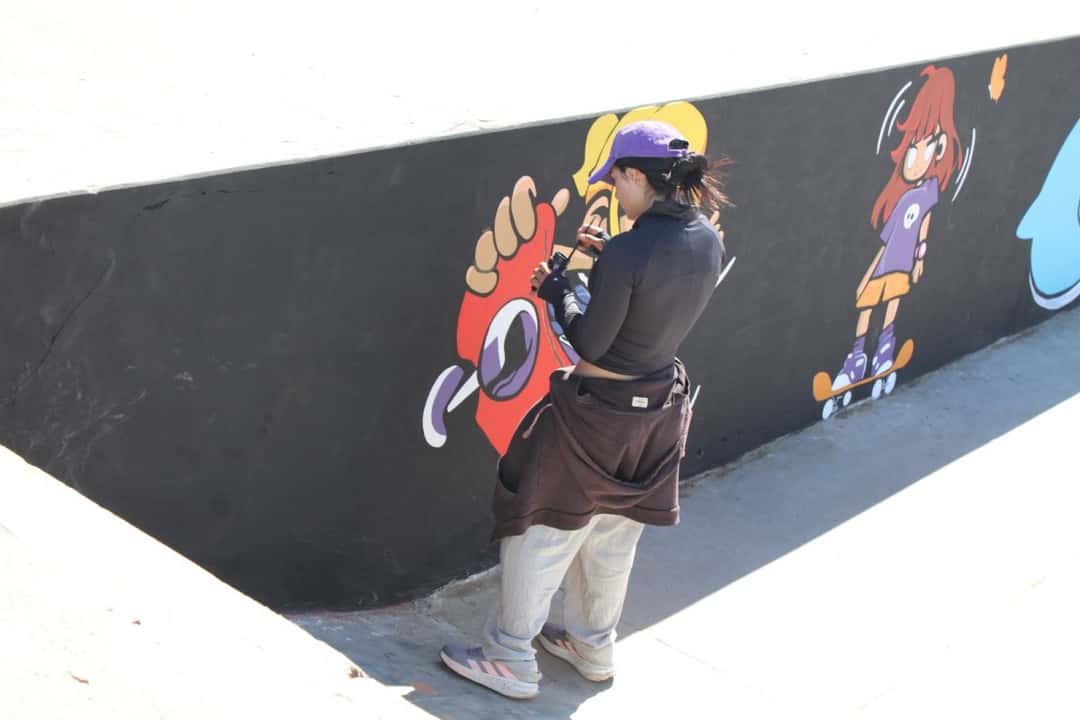 Una artista pinta, sobre una pared negra del skatepark, personajes dibujados en skate y rollers