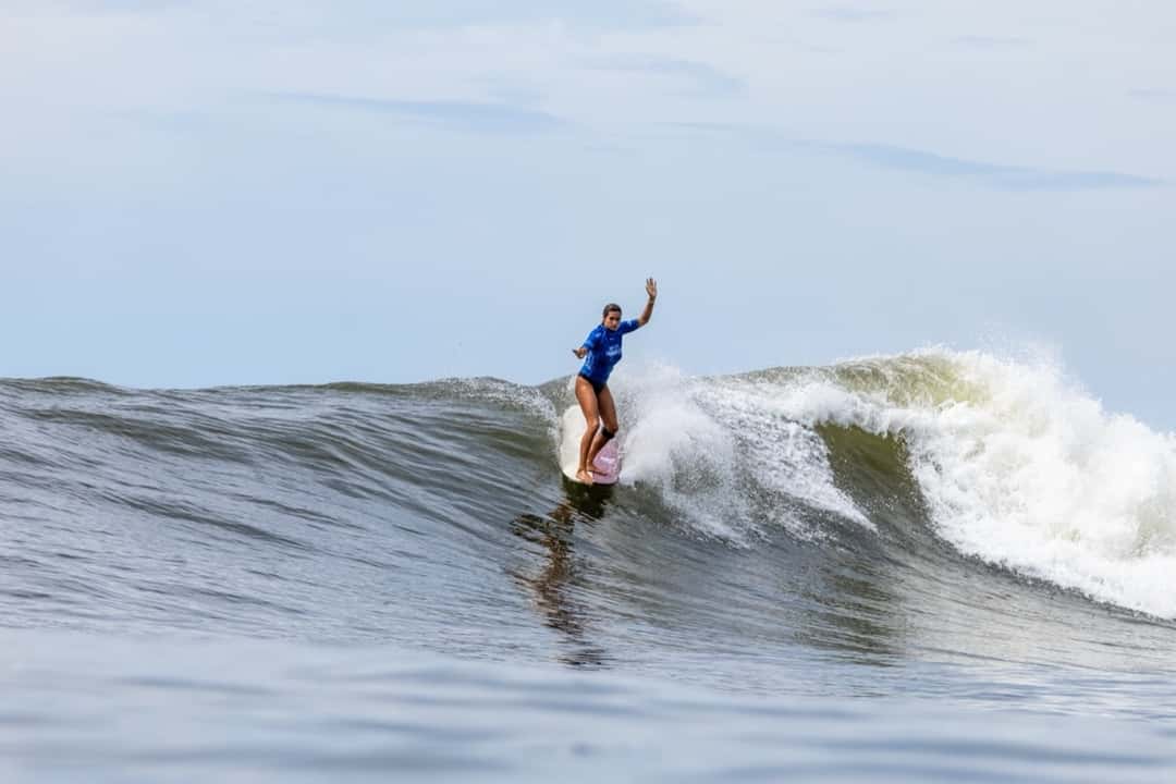 Avalon Gall, con lycra azul, camina sobre su longboard con un brazo en alto en una ola de El Sunzal