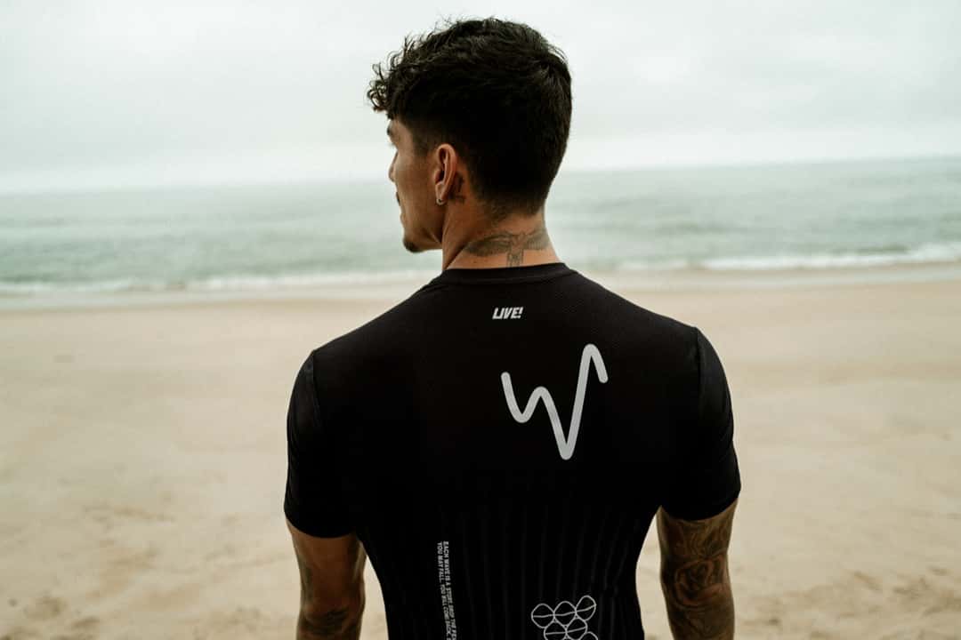 Gabriel Medina, de espaldas en la playa, con la remera negra de la colección: una M blanca con forma de onda en la espalda y, abajo, un dibujo de arcos superpuestos