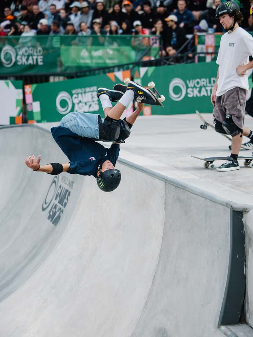 Egoitz Bijueska, con casco negro y buzo azul, gira cabeza abajo sobre el borde del bowl agarrando la tabla, con el público y los carteles de World Skate de fondo