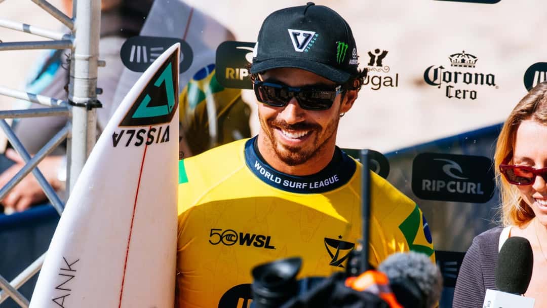 Yago Dora, con gorra negra, anteojos oscuros y la lycra amarilla de líder del ranking con la bandera de Brasil, sonríe con su tabla blanca en Supertubos