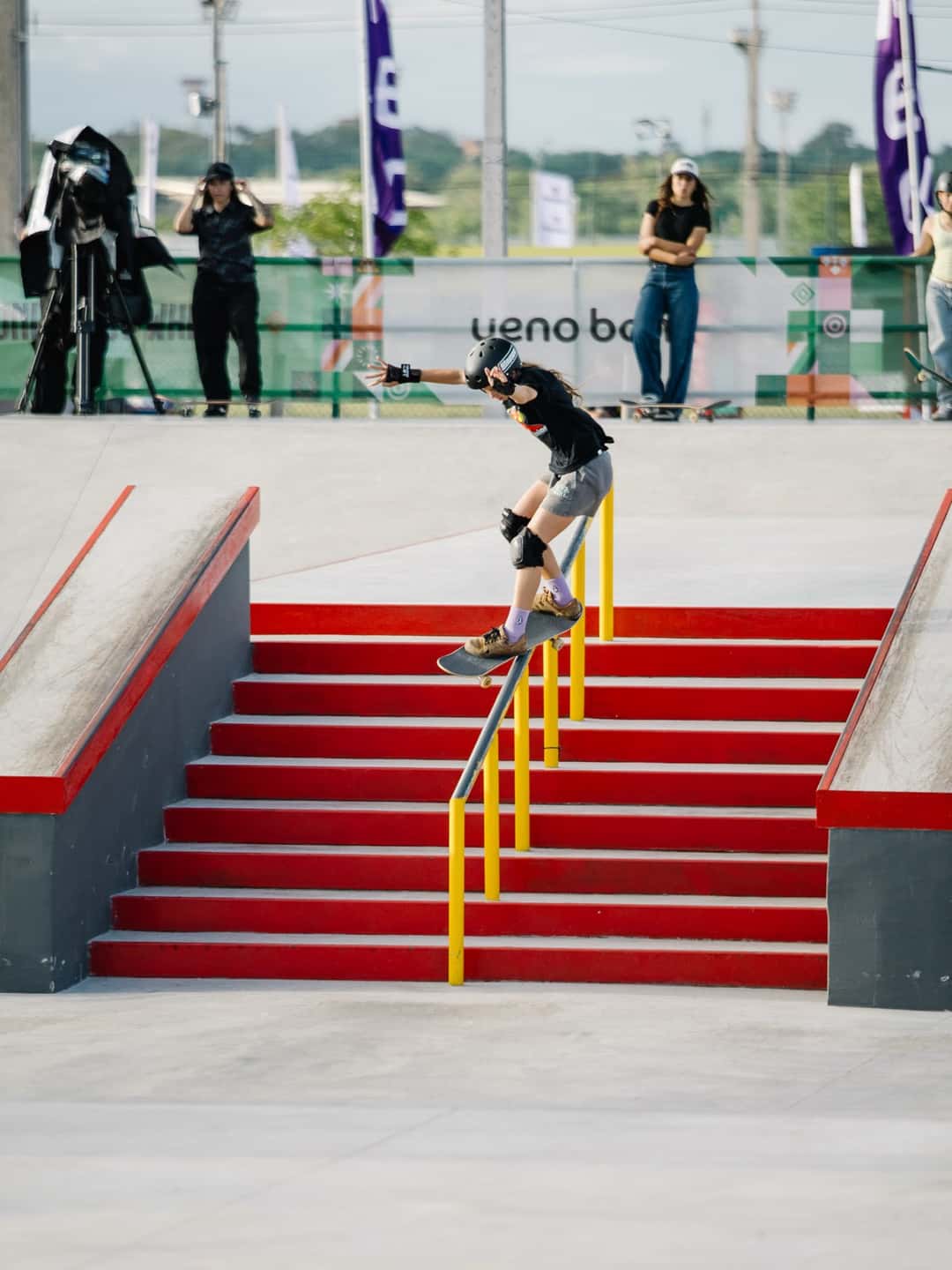 Cora Wilton se desliza sobre la baranda amarilla de la escalera roja de la pista de street de Luque, con cámaras y skaters mirando detrás de la valla