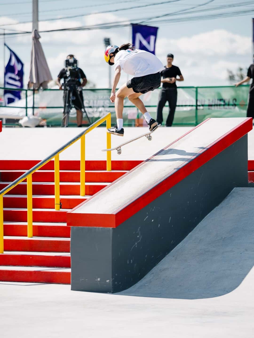 Ginwoo Onodera, con casco y remera blanca, desliza sobre la baranda amarilla de la escalera roja de la pista de street de Luque, con un camarógrafo detrás de la valla