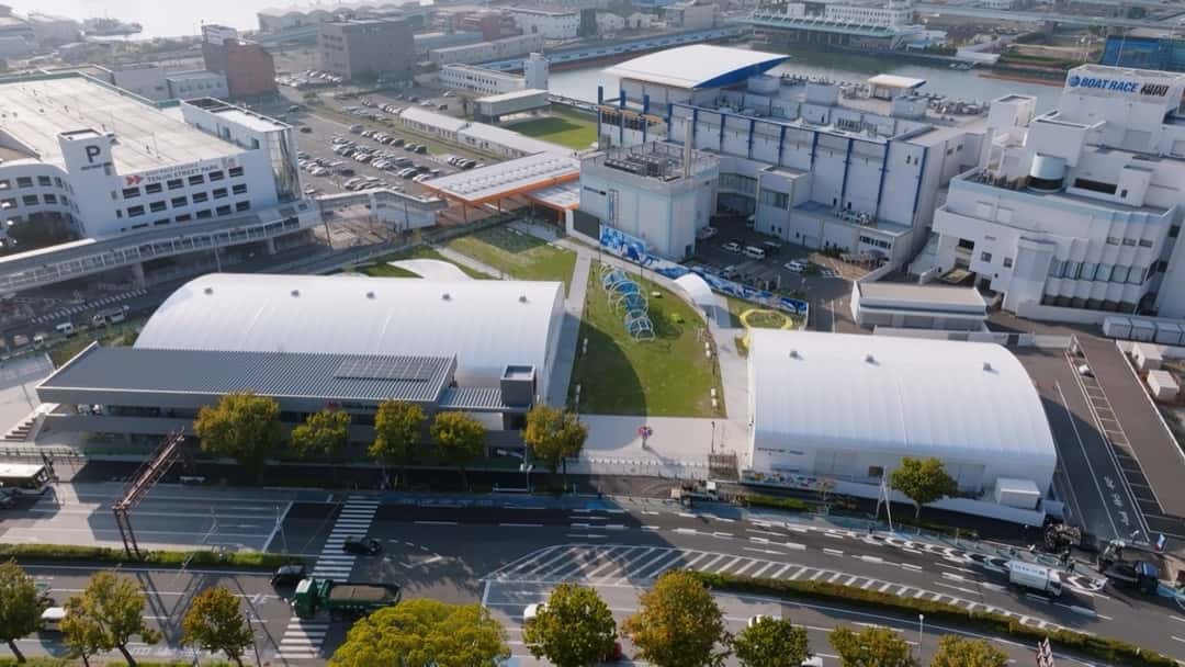 Vista aérea del Tenjin Street Park: dos naves blancas con techo de arco, una plaza de césped en el medio y, al lado, el estadio de carreras de lanchas Boat Race Fukuoka