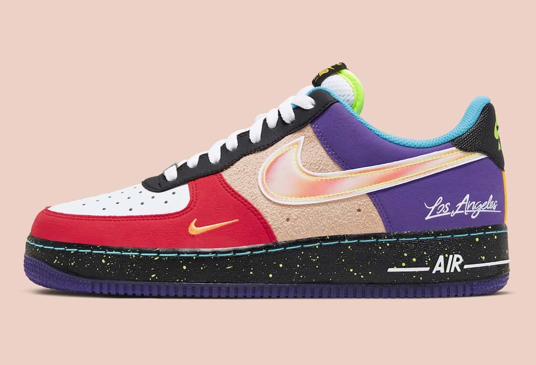 Zapatilla izquierda del Nike Air Force 1 Low What The LA de perfil sobre fondo rosa claro, con puntera blanca, panel rojo con swoosh chico naranja, gamuza beige con un swoosh iridiscente de borde amarillo flúor, talón violeta con la firma Los Angeles en blanco y mediasuela negra con salpicado verde flúor