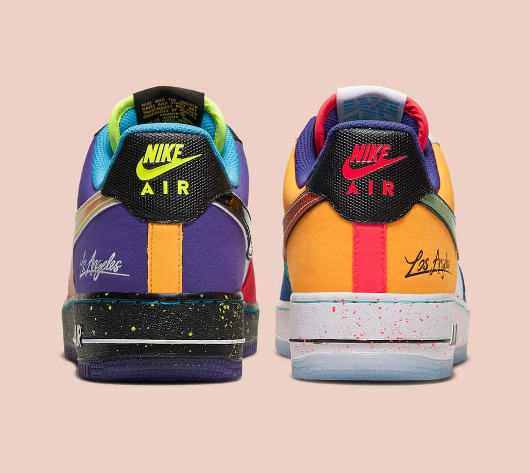 Vista trasera del par del Nike Air Force 1 Low What The LA sobre fondo rosa claro: a la izquierda el talón violeta con una franja naranja, logo Nike Air amarillo flúor, firma Los Angeles en blanco y mediasuela negra salpicada; a la derecha el talón dorado con franja roja, logo Nike Air rojo, firma Los Angeles en negro, mediasuela blanca con salpicado rojo y suela translúcida
