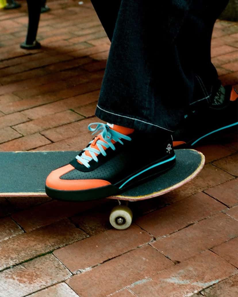 Foto de campaña del BBC ICECREAM x Reebok Board Flip Pumpkin: una persona con jean oscuro ancho apoya el pie sobre una tabla de skate en una vereda de ladrillos, con la zapatilla negra, naranja y celeste