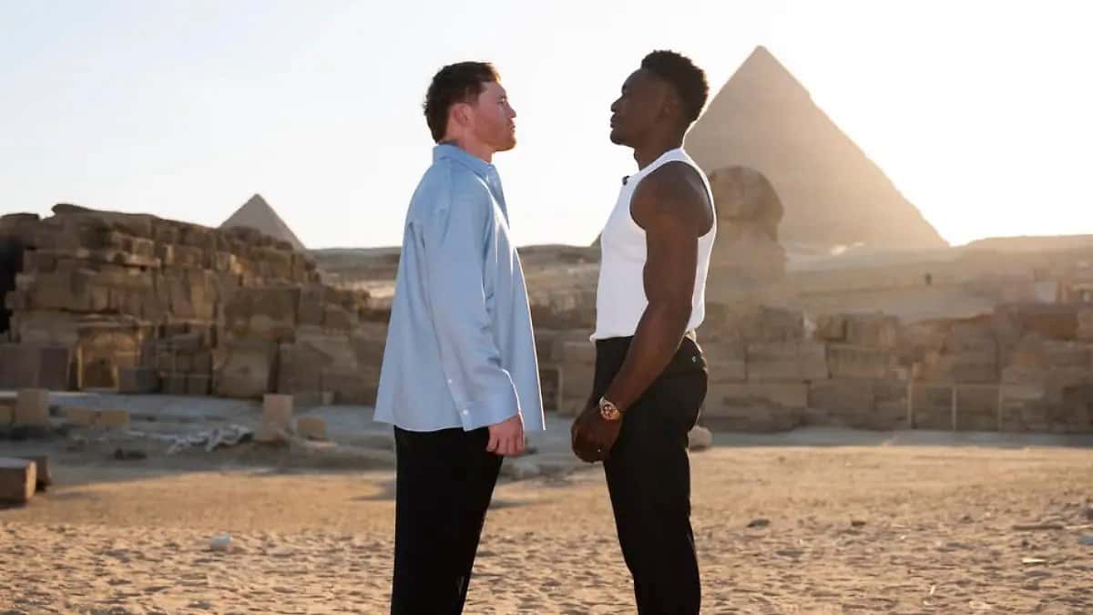 Canelo Álvarez, de camisa celeste, y Christian Mbilli, de musculosa blanca, cara a cara en el desierto con las pirámides de Egipto detrás