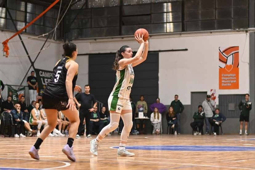 Una jugadora de Ferro, con camiseta blanca y verde, lanza al aro ante la mirada de una rival de negro, en una cancha con público