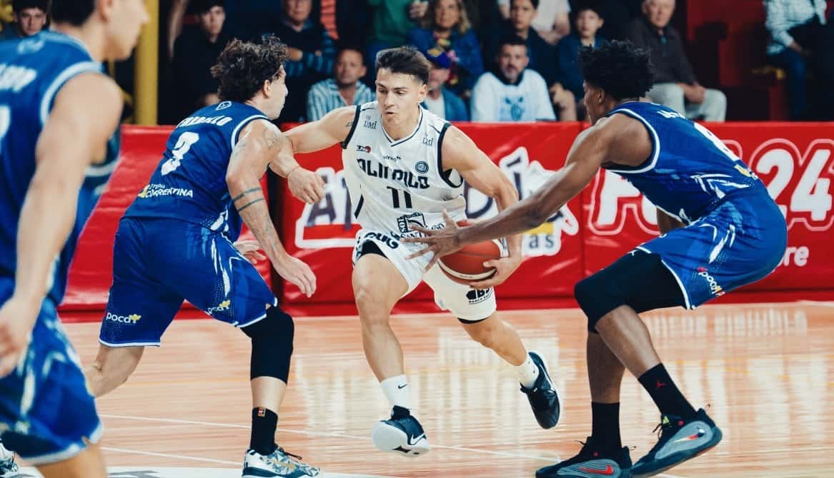 Un jugador de Olímpico de La Banda, con la camiseta blanca número 11, avanza con la pelota entre dos jugadores de Regatas Corrientes vestidos de azul, con la tribuna detrás
