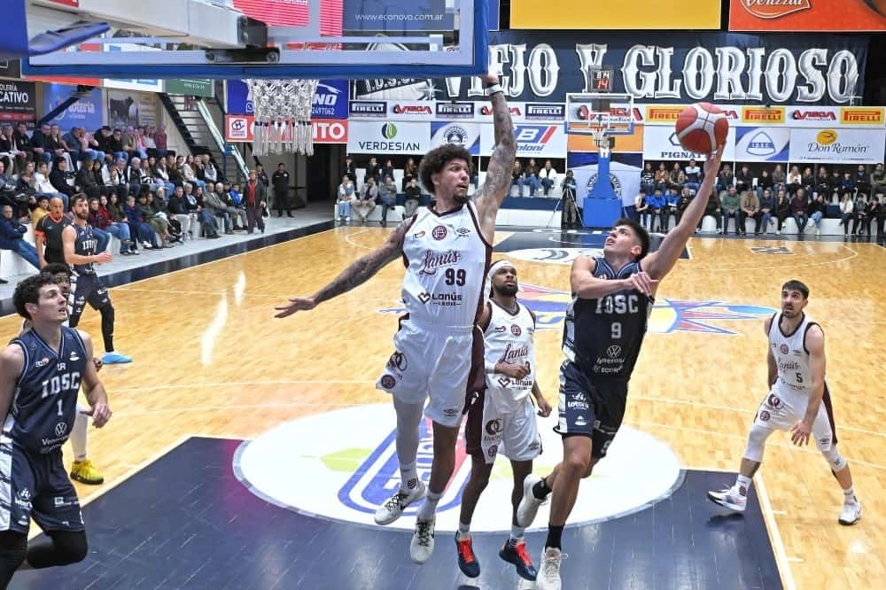 Un jugador de Independiente de Oliva, con camiseta azul oscuro, salta para tirar al aro entre jugadores de Lanús, de blanco, en El Gigante de la Ruta 9