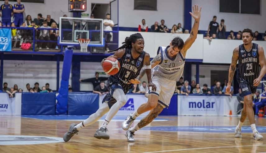 Un jugador de Argentino de Junín, con la camiseta azul oscuro número 3, avanza con la pelota mientras un jugador de Boca, de blanco, levanta el brazo para defenderlo en El Fortín de las Morochas