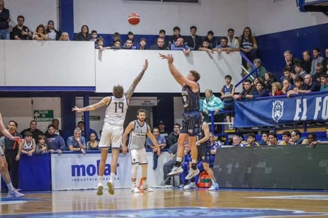 Un jugador de Argentino de Junín, de azul oscuro, lanza en suspensión ante la marca de un jugador de Boca, de blanco, con la tribuna local de fondo