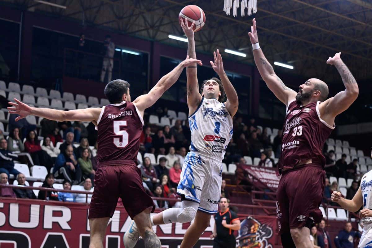 Un jugador de Regatas Corrientes, de blanco y celeste, lanza en suspensión entre dos jugadores de Lanús, de granate, en el estadio Antonio Rotili