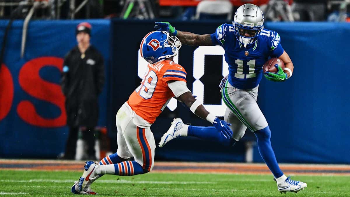Jaxon Smith-Njigba, con la camiseta azul número 11 de los Seahawks, corre con la pelota y aparta con el brazo a Ja'Quan McMillian, número 29 de los Broncos, en el Empower Field at Mile High