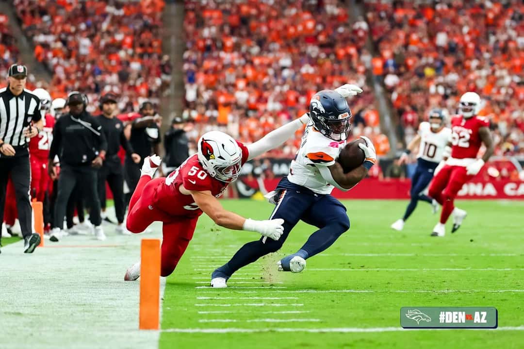 RJ Harvey, de los Broncos, con la camiseta blanca, se tira hacia adelante con la pelota mientras un defensor de los Cardinals intenta tacklearlo junto a la línea lateral