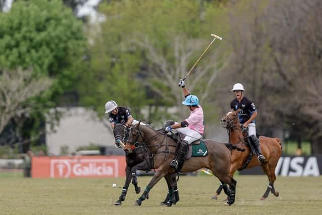 Un jugador de La Ensenada, con camiseta rosa y casco celeste, levanta el taco para pegarle a la bocha entre dos jugadores de Don Ercole de camiseta azul oscuro, con carteles de los sponsors del torneo de fondo