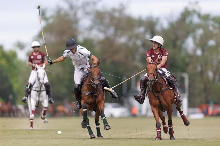 Un jugador de camiseta blanca le pega a la bocha con el taco, perseguido por dos rivales de camiseta bordó, en una cancha del Tortugas Country Club