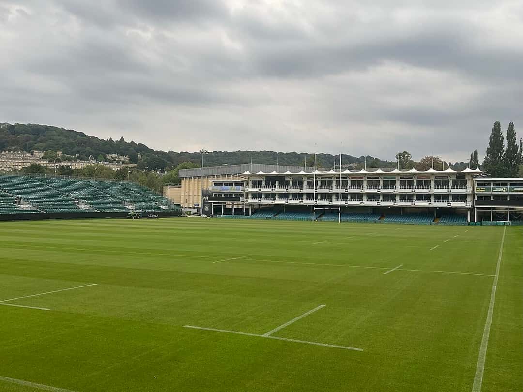 Imagen del estadio: la cancha del Recreation Ground de Bath, con el césped verde y la tribuna principal al fondo bajo un cielo nublado