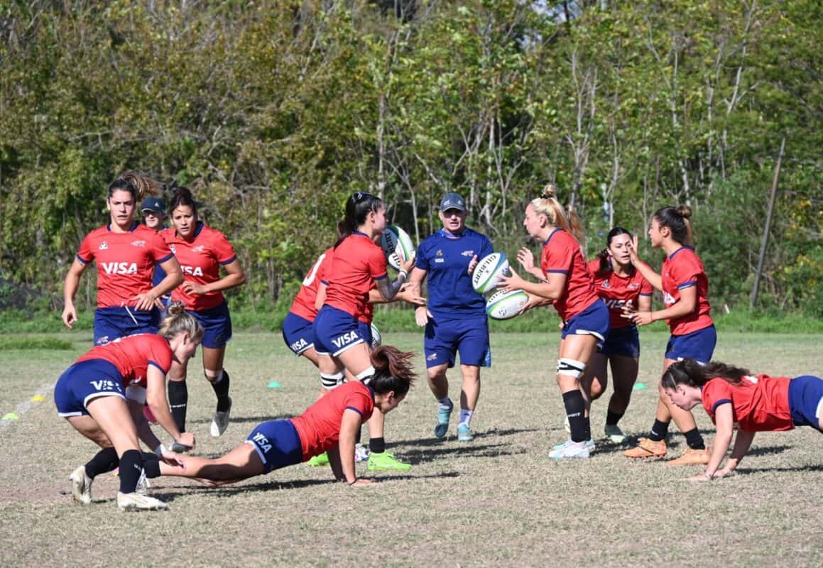Foto de archivo: jugadoras de rugby con camiseta roja y short azul hacen un ejercicio de entrenamiento junto a un entrenador, en una cancha de pasto