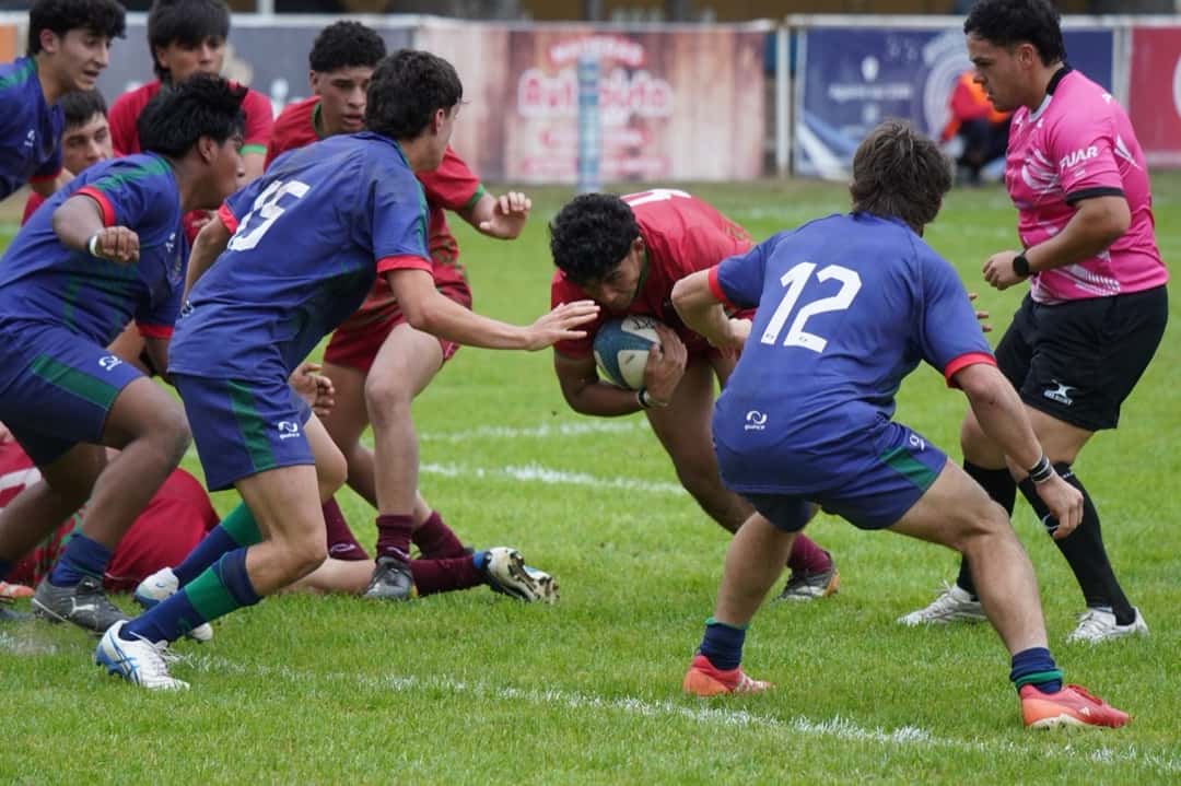 Juveniles de los seleccionados de Andina y Jujeña durante su partido del Select 12 Norte: un jugador de camiseta roja avanza con la pelota entre rivales de azul, con el árbitro de rosa a la derecha