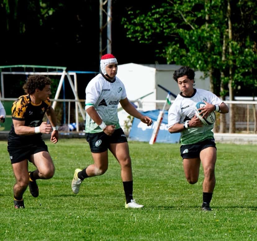 Foto de archivo: tres jugadores juveniles corren en un partido de rugby: uno de camiseta blanca y verde lleva la pelota, seguido por un compañero con casco rojo y un rival de negro y amarillo
