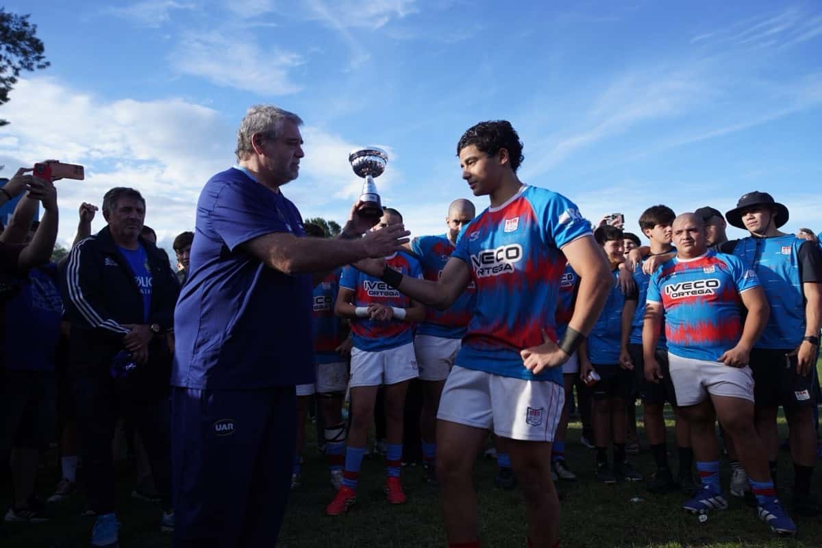 Un jugador del seleccionado juvenil de la Unión Santiagueña, con camiseta celeste y roja, recibe la copa de campeón del Select 12 Norte de manos de un hombre con ropa de la UAR, con sus compañeros detrás
