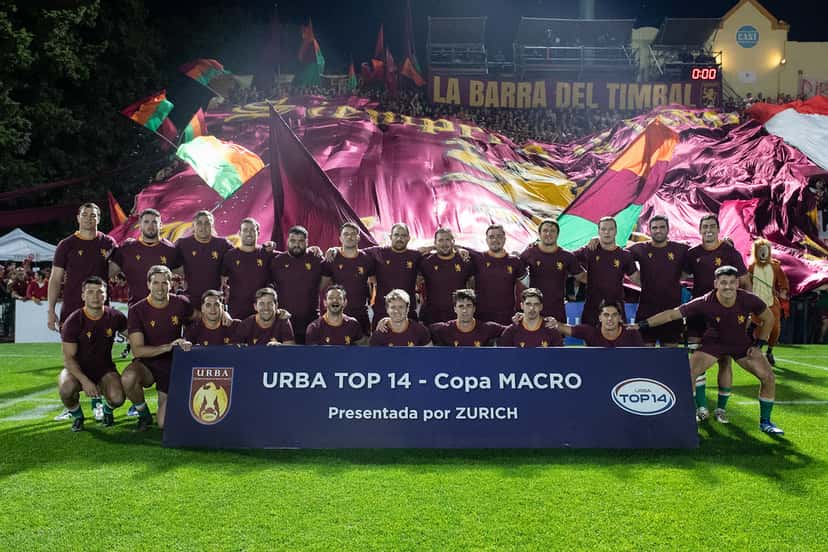 El plantel de Newman, con la camiseta bordó, posa en la cancha del CASI antes de la semifinal, delante de un cartel del URBA Top 14 y con banderas en la tribuna