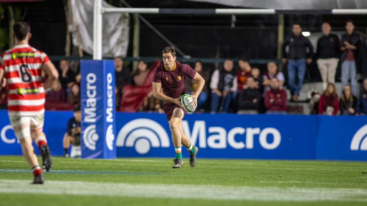 Un jugador de Newman, con la camiseta bordó, corre con la pelota en la mano con un rival de Alumni, de rojo y blanco, en primer plano, en la semifinal del URBA Top 14