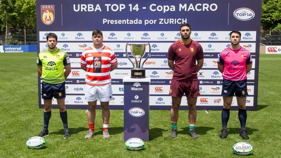 Un jugador de Alumni, con camiseta a rayas rojas y blancas, y uno de Newman, de bordó, posan a los costados de la copa del URBA Top 14, frente a un panel con la leyenda «URBA Top 14 - Copa Macro»