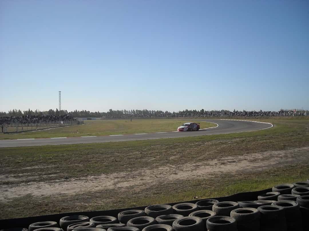 Foto de archivo: un auto de carrera rojo y blanco dobla una curva del autódromo de Rosario, con público en las tribunas del fondo y una barrera de cubiertas adelante